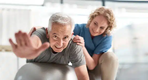 male senior stretching while on an exercise ball supported by a female therapist