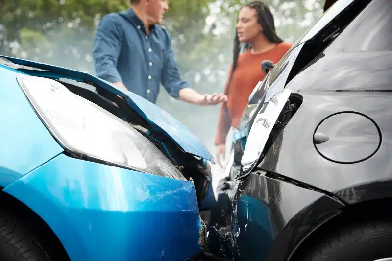 close up of two car bumpers that crashed with two owner's having an argument in the background